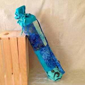 May include: A turquoise yoga mat carrier with a patchwork design featuring blue and white floral patterns. The carrier has a drawstring closure and is made of fabric.