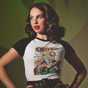 May include: A white and black baseball tee with the text "Creep Tease" and a mummy graphic. The model is wearing black shorts and fishnet stockings. The background is a solid green colour.