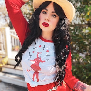 Może przedstawiać: Kobieta w czerwono-białej koszulce bejsbolowej z grafiką diablicy żonglującej nożami i sercami. Ma na sobie szeroki słomkowy kapelusz i czerwoną spódnicę ze złotym paskiem w kształcie serca.