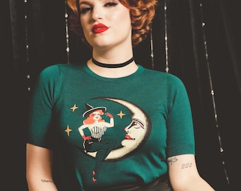 Puede incluir: Camiseta verde de manga corta con un dibujo de una bruja en una luna creciente. La bruja lleva un sombrero y guantes rojos. La persona sostiene una calabaza a rayas blancas y negras. Hay otras calabazas apiladas cerca.