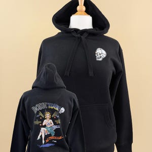 Pode incluir: Uma camisola preta com capuz apresentando um gr&aacute;fico de caveira branca no peito esquerdo. As costas exibem uma ilustra&ccedil;&atilde;o colorida de uma figura loira com chap&eacute;u e saia, com o texto "BONE TEASE" e "Born to Be Wild!!" impresso acima e abaixo.