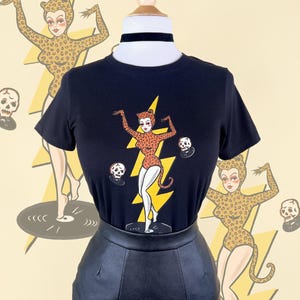 Może przedstawiać: Czarny T-shirt z grafiką przedstawiającą kobietę w stroju w panterkę tańczącą na gramofonie z czaszkami. Kobieta trzyma błyskawicę. Na koszulce nadrukowany jest napis "Rockin' Dead".