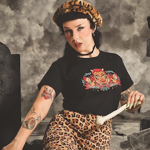 May include: A black short-sleeved t-shirt featuring a graphic print of a red devil face with the text LOST SOUL, paired with leopard print trousers and a matching leopard print beret. The shirt has a crew neckline and a central colourful illustration.