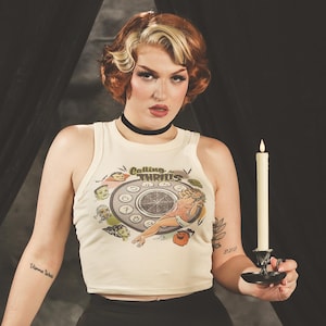 May include: A cream-coloured sleeveless crop top featuring a graphic print of a rotary telephone with the text "Calling for THRILLS" surrounded by various monster illustrations, paired with a black choker and solid black high-waisted trousers.