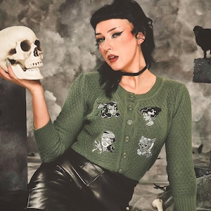 May include: An olive green button-down cardigan with three-quarter sleeves and a textured knit pattern. The front features four embroidered patches depicting stylised monster characters. The cardigan has a round neckline and matching green buttons.