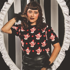 May include: A black short-sleeved knit jumper featuring a repeating pattern of red and white clown faces and red dots. The jumper is worn with a black skirt and black sheer wrist cuffs, framed by an oval border with white ruffles and pearls.