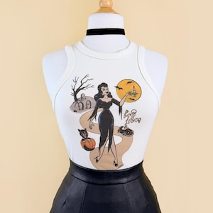 Peut inclure: Un débardeur blanc sans manches avec une impression graphique représentant une personne en robe noire sur un chemin avec une citrouille, un chat noir, un fantôme, une voiture, un arbre dénudé, des pierres tombales et une lune jaune, avec un ras-de-cou noir.
