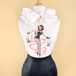 May include: White tank top featuring a vintage-style illustration of a woman holding clown dolls, worn with a black pencil skirt and a large white ruffled collar. The mannequin is set against a pale yellow backdrop.