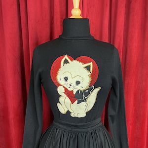 May include: Black turtleneck sweater featuring a white cat holding a red heart graphic. The cat is wearing a black bow tie. The sweater is paired with a black full skirt.