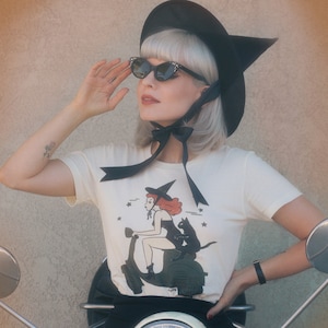 May include: A woman wearing a black witch hat and sunglasses sits on a white scooter. She is wearing a white t-shirt with a black bow and a black and white illustration of a witch riding a scooter with a black cat.