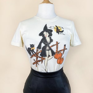Może przedstawiać: Biały t-shirt z vintage'ową ilustracją czarownicy z czarnym kotem i dyniami. Czarownica ma na sobie czarną sukienkę i spiczasty kapelusz. Ilustracja jest w stylu kreskówkowym.