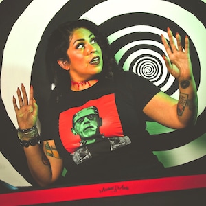 May include: Black t-shirt with a red square featuring a green and black Frankenstein's monster graphic. The background has a black and white spiral design. The person is wearing bracelets and has tattoos.