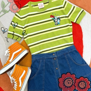 May include: A lime green striped top with a cartoon character, a denim skirt, and orange high-top trainers. The top has black and white stripes. The skirt has flower appliqués. The trainers are orange.