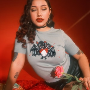 May include: A pale blue short-sleeved top with a bat design, red bow, and heart details. The top is worn with black shorts. A pink rose is held in the hand.