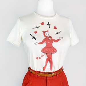 Może przedstawiać: Kremowy t-shirt z czerwoną grafiką diabła żonglującego nożami i sercami. Koszulka jest połączona z czerwonymi spodniami i brązowym paskiem. Strój jest prezentowany na manekinie.
