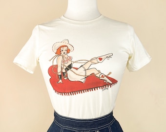 Può includere: Una maglietta bianca con una stampa grafica di una donna con un cappello da cowboy e stivali, seduta su un tappeto rosso a forma di cuore. La donna indossa una camicetta a quadri rossi e bianchi e una cintura nera.