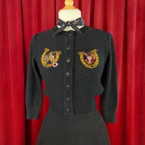 Pode incluir: Cardigan preto de malha texturizada com ferraduras bordadas. Uma ferradura apresenta a cabeça de um cavalo e um coração vermelho, a outra apresenta uma mulher com um chapéu de cowboy.