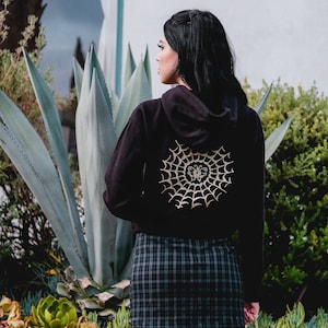 May include: Black hoodie with a gold spiderweb and butterfly design on the back. The hoodie is worn by a person with tattoos on their legs.