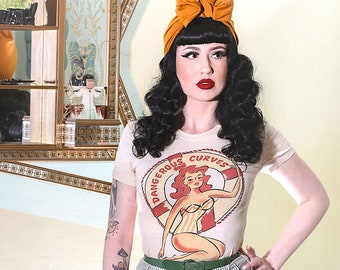Può includere: Una donna indossa una maglietta bianca con una grafica di una donna in un salvagente e il testo "Dangerous Curves". Indossa anche una cintura verde, una gonna a righe e una borsa di vimini marrone. Ha un grande fiocco arancione nei capelli.