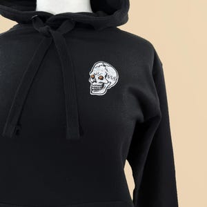 Pode incluir: Uma camisola preta com capuz apresentando um gr&aacute;fico de caveira bordado em branco na parte superior esquerda do peito. O capuz inclui um cord&atilde;o preto a condizer no decote e um bolso frontal tipo canguru, exibido num manequim.