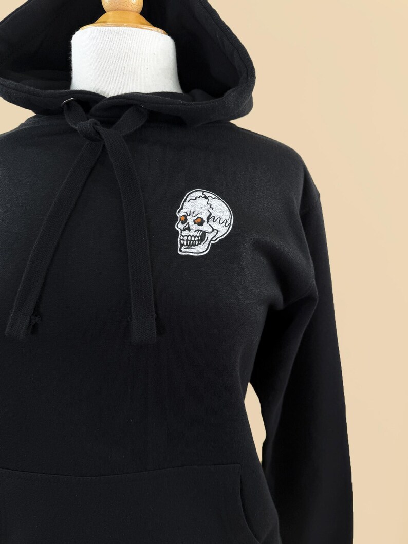 Pode incluir: Uma camisola preta com capuz apresentando um gr&aacute;fico de caveira bordado em branco na parte superior esquerda do peito. O capuz inclui um cord&atilde;o preto a condizer no decote e um bolso frontal tipo canguru, exibido num manequim.