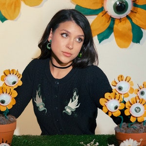May include: A woman with dark hair wearing a black cardigan with embroidered hands and snakes. She is standing in front of a white wall with yellow and green paper flowers with googly eyes.