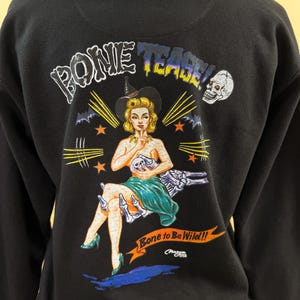 Pode incluir: Uma camisola preta com capuz apresentando uma estampa gr&aacute;fica nas costas. O design exibe uma figura estilo pin-up com chap&eacute;u de bruxa, um cr&acirc;nio e o texto "BONE TEASE!" e "Bone to Be Wild!!" com m&atilde;os esquel&eacute;ticas e estrelas laranja.