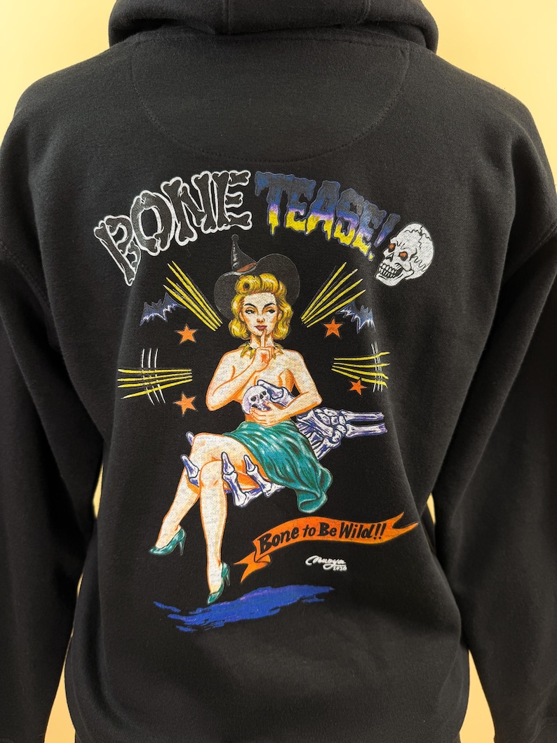 Pode incluir: Uma camisola preta com capuz apresentando uma estampa gr&aacute;fica nas costas. O design exibe uma figura estilo pin-up com chap&eacute;u de bruxa, um cr&acirc;nio e o texto "BONE TEASE!" e "Bone to Be Wild!!" com m&atilde;os esquel&eacute;ticas e estrelas laranja.