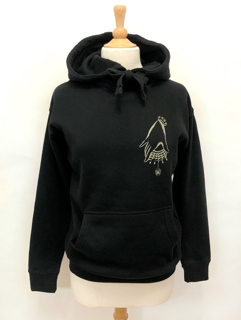 May include: Black hooded sweatshirt with a gold embroidered design of a hand with a spider web and a spider on the left chest.
