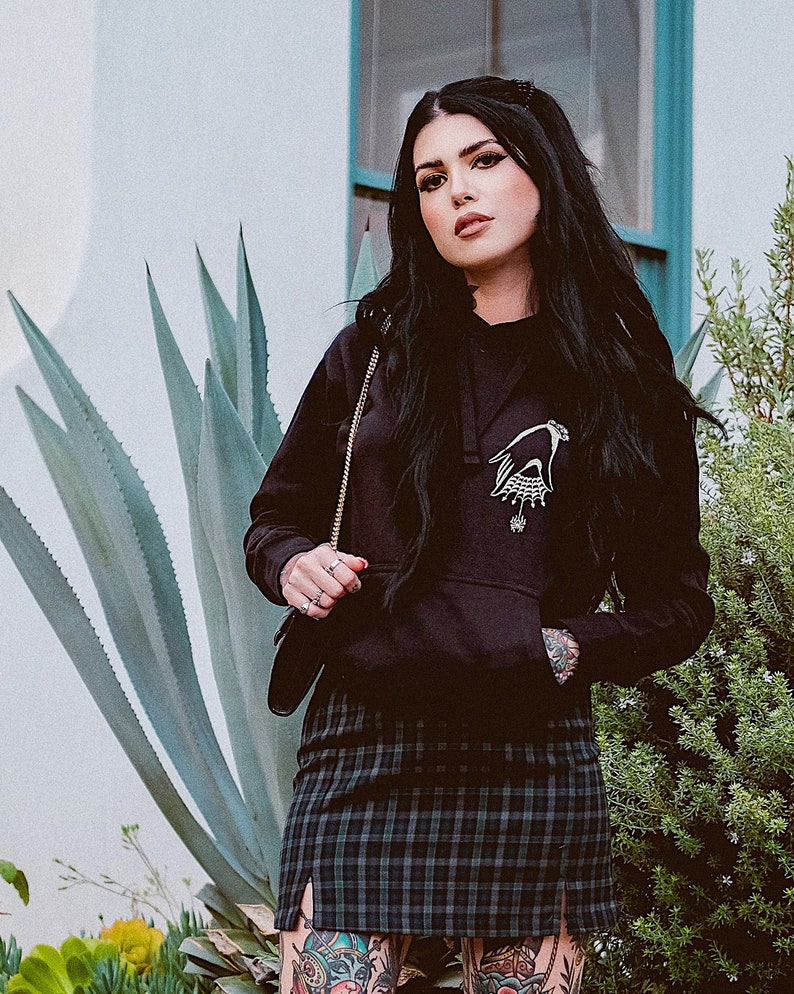 May include: A black hoodie with a white graphic of a hand holding a spider web. The hoodie is worn by a person with black hair and tattoos. The person is wearing a black and green plaid skirt.