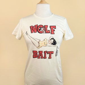 Pu&ograve; includere: Una maglietta bianca con una grafica rossa e nera di una donna in costume da bagno con una testa di lupo sul petto. Il testo "WOLF BAIT" &egrave; stampato in lettere rosse sotto la grafica.