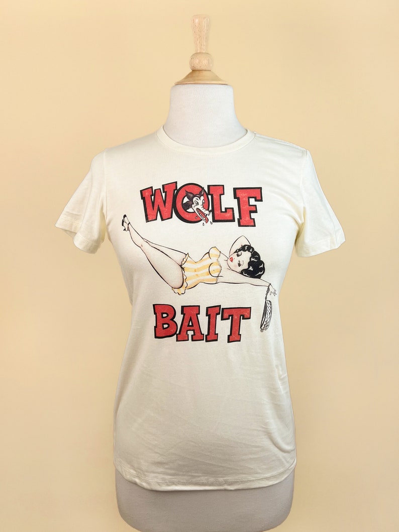 Pu&ograve; includere: Una maglietta bianca con una grafica rossa e nera di una donna in costume da bagno con una testa di lupo sul petto. Il testo "WOLF BAIT" &egrave; stampato in lettere rosse sotto la grafica.