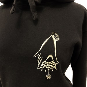 May include: Black hoodie with a gold embroidered design of a hand holding a spider web with a spider in the center.