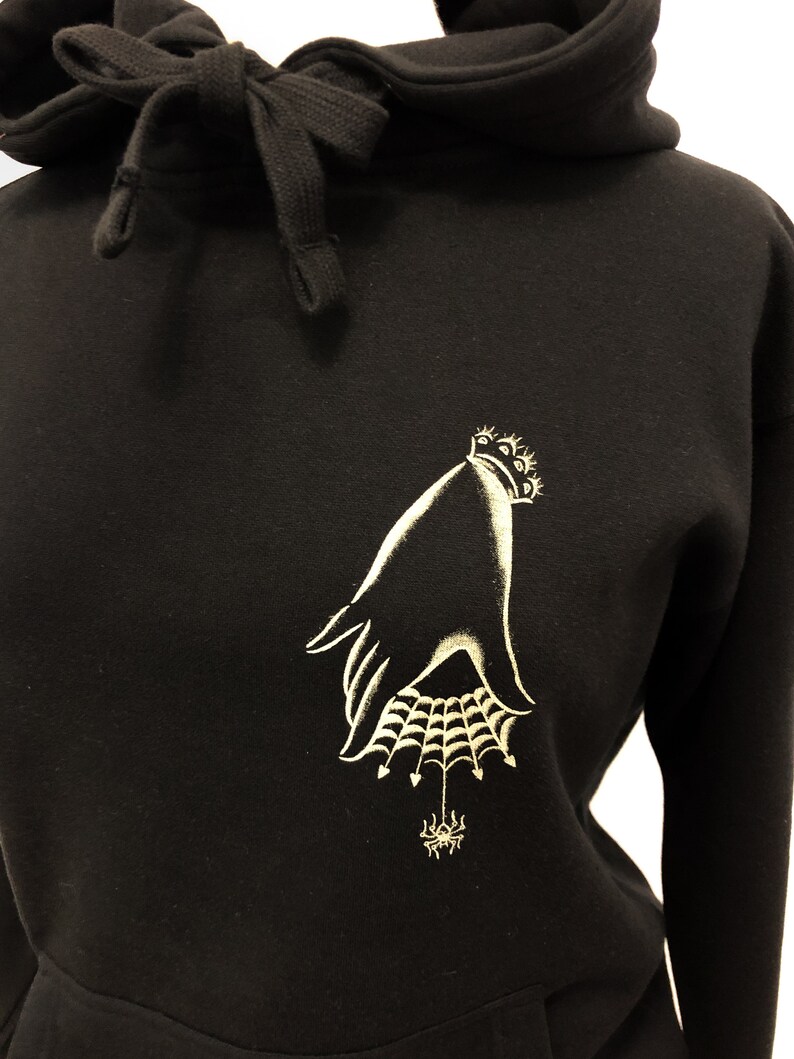 May include: Black hoodie with a gold embroidered design of a hand holding a spider web with a spider in the center.