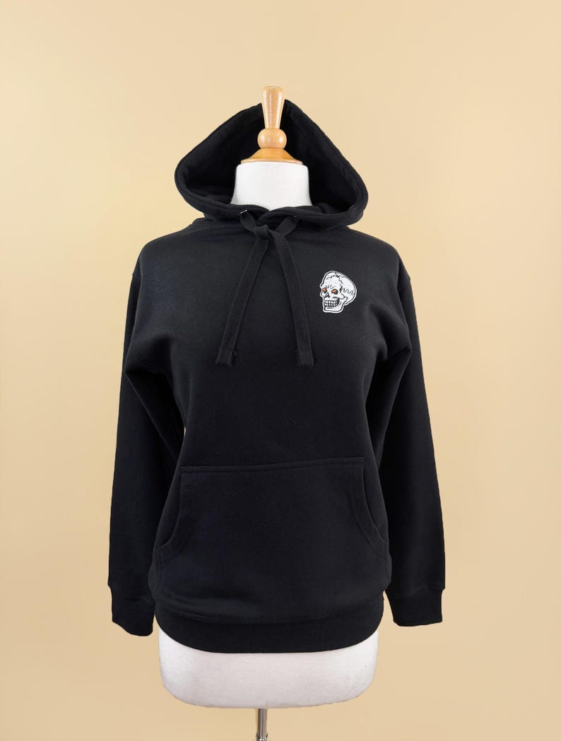Pode incluir: Uma camisola preta de manga comprida com capuz, apresentando um gr&aacute;fico de caveira branca na parte superior esquerda do peito. O capuz inclui um bolso canguru, cord&otilde;es pretos a condizer e punhos canelados nas mangas e na bainha inferior.