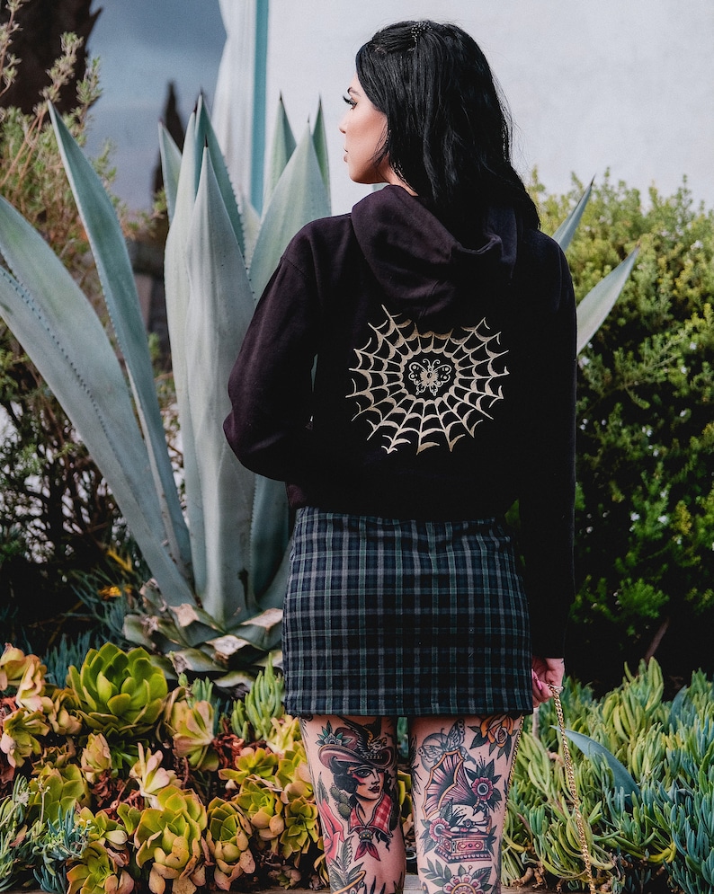 May include: Black hoodie with a gold spiderweb and butterfly design on the back. The hoodie is worn by a person with tattoos on their legs.