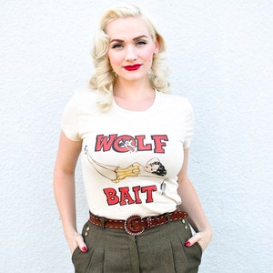 Pu&ograve; includere: Una donna con i capelli biondi e il rossetto rosso indossa una maglietta bianca con una grafica rossa e nera che dice "WOLF BAIT". Indossa anche pantaloni marroni con una cintura marrone.