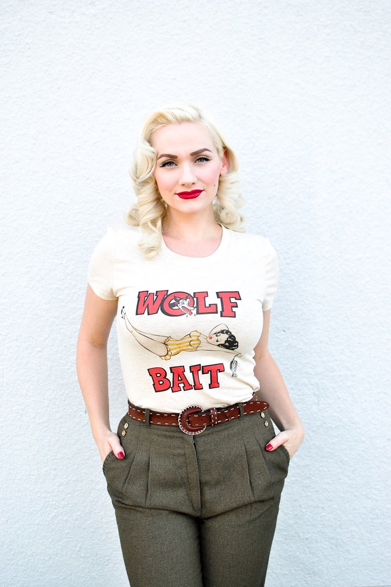 Pu&ograve; includere: Una donna con i capelli biondi e il rossetto rosso indossa una maglietta bianca con una grafica rossa e nera che dice "WOLF BAIT". Indossa anche pantaloni marroni con una cintura marrone.