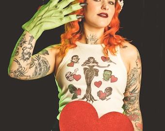 May include: A woman with red hair and tattoos is holding a large red heart. She is wearing a white tank top with a print of a monster and a woman. She is also wearing green gloves with long claws.
