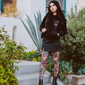 May include: A woman wearing a black hoodie with a graphic print, a plaid skirt, and black boots. She has tattoos on her legs.