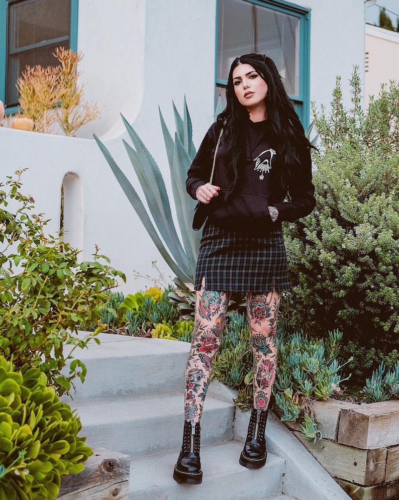 May include: A woman wearing a black hoodie with a graphic print, a plaid skirt, and black boots. She has tattoos on her legs.