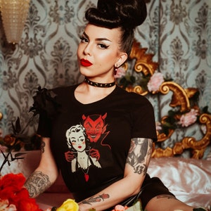 Op de afbeelding: Een vrouw met donker haar dat in een hoge knot is gemodelleerd, draagt een zwart T-shirt met een afbeelding van een vrouw en een duivel. Ze zit op een bed met rode en gele bloemen.