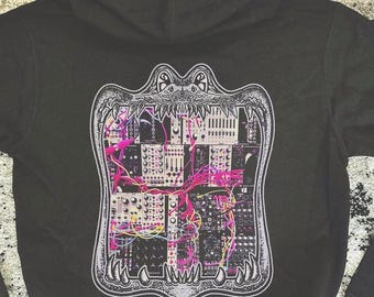 May include: A black hooded sweatshirt featuring a large graphic on the back. The design depicts a stylized creature with an open mouth, framing a detailed illustration of modular synthesizer equipment with bright pink and yellow patch cables.