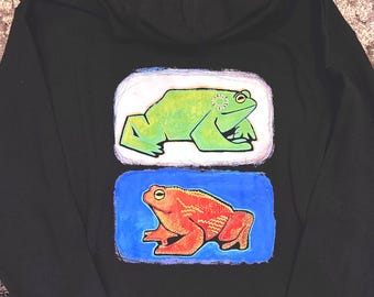 May include: A black hooded sweatshirt featuring a graphic print on the back. The design consists of two rectangular panels, one above the other. The top panel shows a green frog on a white background, and the bottom panel shows an orange frog on a blue background.