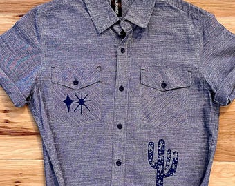 May include: A short-sleeved button-down shirt in a heathered blue fabric. The shirt features a pointed collar, two chest pockets, and a central button placket. Dark blue graphics include a starburst symbol and a cactus on the front.