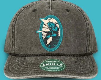 May include: A charcoal gray baseball cap with a rope detail and a curved brim. The cap features an oval patch with a bear graphic in teal and white. The text "LEGACY SKULLY" is on a green sticker on the brim.