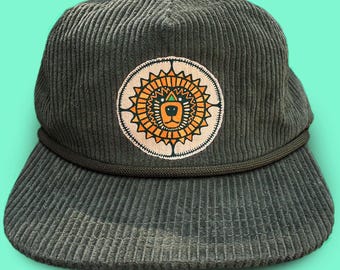 May include: A dark green corduroy baseball cap with a circular patch featuring a lion design in orange, yellow, and green. The cap has a curved brim and a dark green rope detail around the crown.