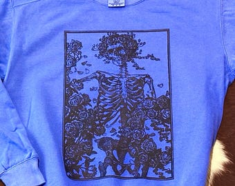 May include: A periwinkle blue long-sleeve crewneck sweatshirt featuring a rectangular black graphic print on the chest. The design depicts a human skeleton surrounded by blooming roses and foliage, enclosed within a thin black rectangular border.