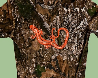 May include: A short-sleeved t-shirt with a camouflage pattern in shades of brown, green, and white. An orange salamander graphic is centered on the front. The shirt is made of a soft material.