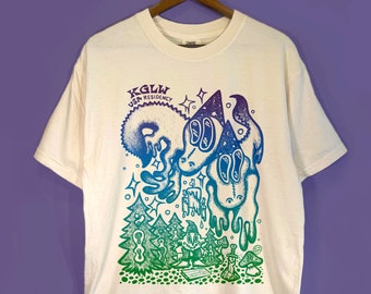 May include: A cream-colored short-sleeve t-shirt featuring a gradient graphic print in purple, blue, and green. The design includes the text KGLW USA RESIDENCY, stylized creatures, pine trees, a wizard figure, a sword, and stars.
